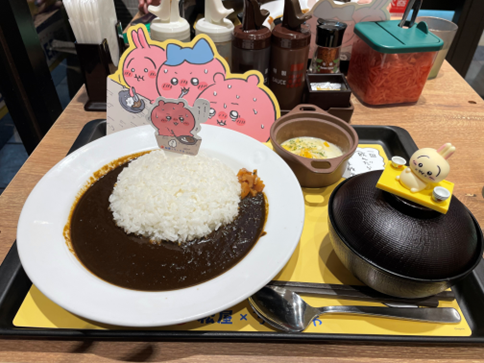 キャラクターのパネルが飾られた黒いカレーライスやお椀などが乗ったコラボカフェのトレイ